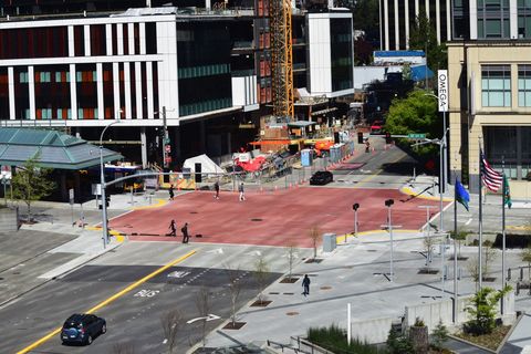 A photo of NE 6th Street and 110th Avenue NE from above. It shows the red patterned concrete of the raised intersection. The transit center is visible on the left and the edge of the light rail station on the right.