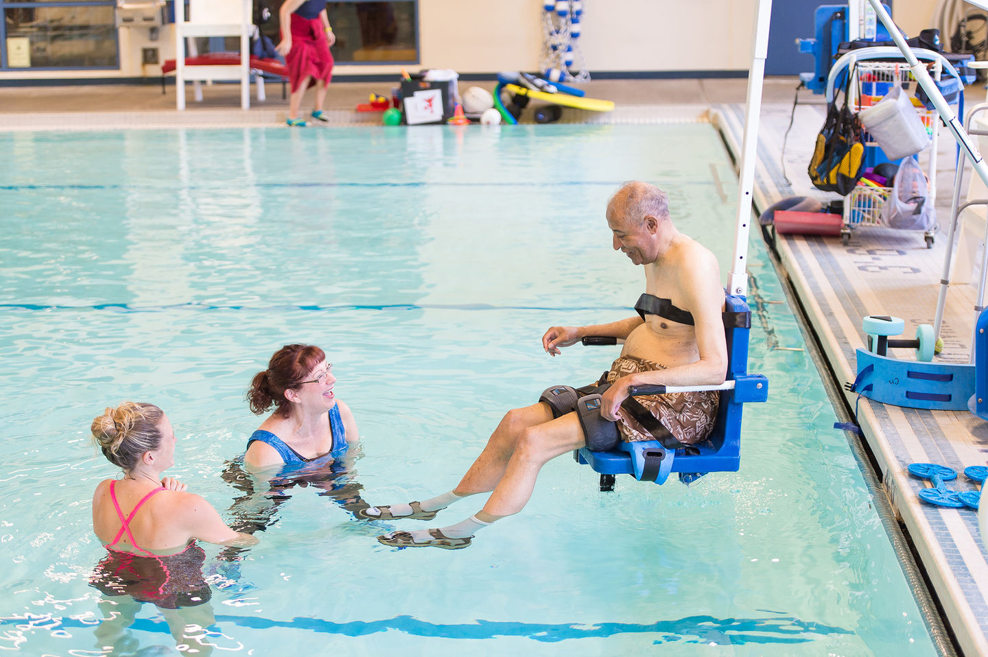 image of three people using a pool. Two women are in the pool, and one man is entering the pool using a pool lift.