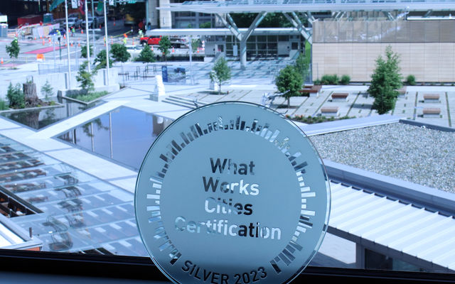 What Works Cities plaque leans against City Hall window, with plaza showing through the glass.