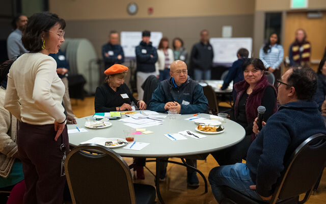 A man speaks into a microphone at a table with several people, in a large room with even more people.