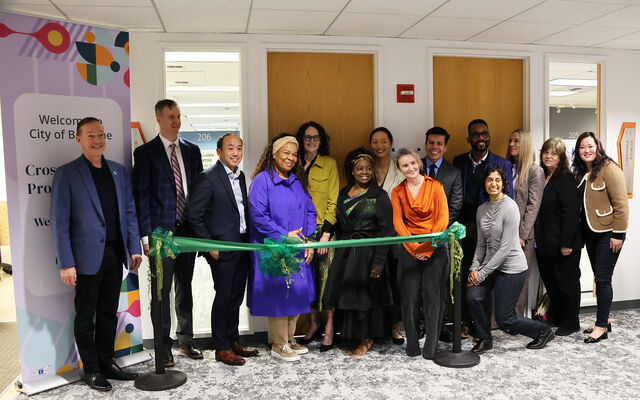 City and school district leaders stand in a room at the Welcome Center behind a green ribbon that they will cut.