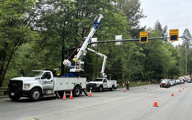 A boom truck and bucket truck with one worker each is erecting a large radar sign pole across a four-lane road. Traffic has been stopped by a uniformed police officer, and traffic cones are in place to divert traffic away from work crews.