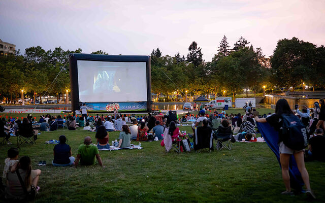People sitting on the lawn at Downtown Park watch a movie on a massive, temporary screen.