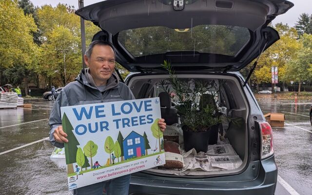 A man holds a sign that says "We love our trees" while he stands in front of his car, which is loaded with free trees.