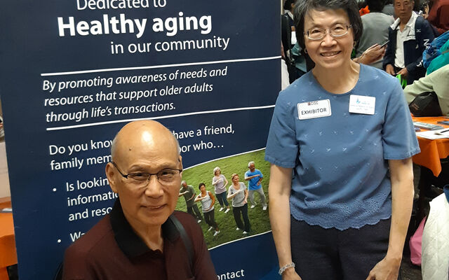 A man and a woman who are members of the Network on Aging stand and sit by a display board about the group.