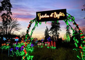 Image shows a lighted arch after sunset with brightly lit floral art in front of, under and around the arch, as well as trees in the backgroun. 