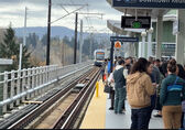 A crowd of people wait at a light rail station as a train arrives on the track.