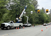 A boom truck and bucket truck with one worker each is erecting a large radar sign pole across a four-lane road. Traffic has been stopped by a uniformed police officer, and traffic cones are in place to divert traffic away from work crews.