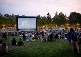 People sitting on the lawn at Downtown Park watch a movie on a massive, temporary screen.