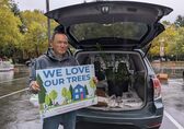A man holds a sign that says "We love our trees" while he stands in front of his car, which is loaded with free trees.