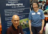 A man and a woman who are members of the Network on Aging stand and sit by a display board about the group.
