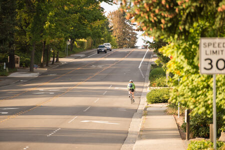 A person bikes away from the viewer and cars are coming towards the camera on a four lane road. In the foreground is a 30 mph speed limit sign.
