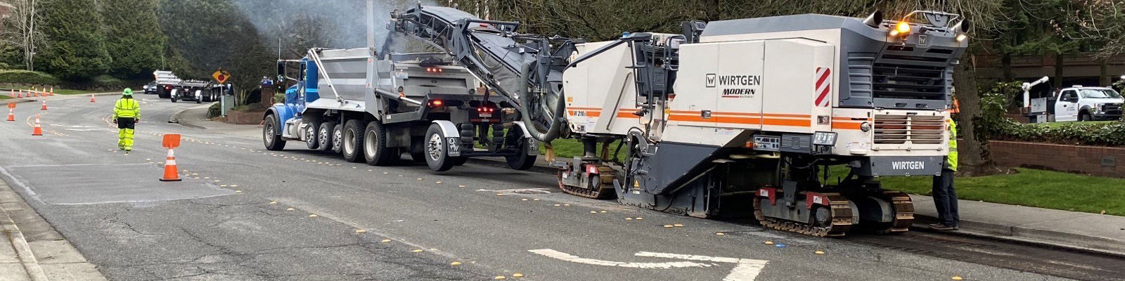 A dump truck works with another machine to scarify the roadway in preparation for repaving. There are construciton cones and two people dressed in high visibility clothes on the left side of the roadway.