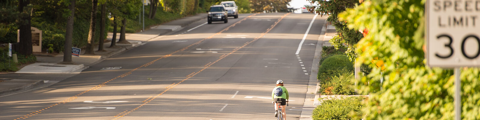 A person bikes away from the viewer and cars are coming towards the camera on a four lane road. In the foreground is a 30 mph speed limit sign.