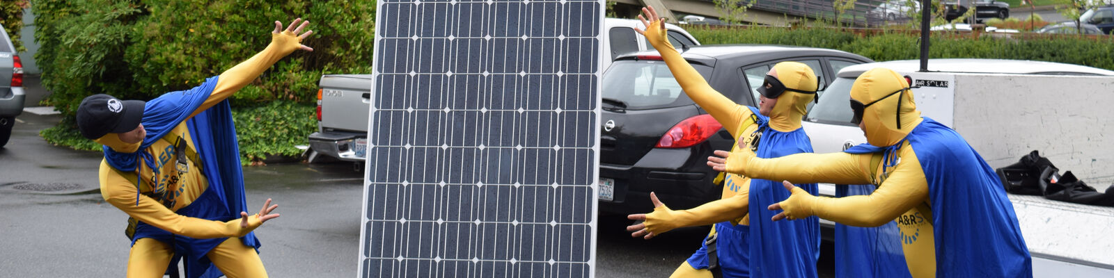 Staff dressed in superhero costumes show off a solar panel in a parking lot.