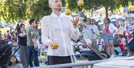 A man juggles three balls at Downtown Park while many people enjoy themselves behind him.