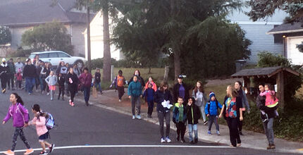 A large group of schoolchildren and parents walk in a procession down a neighborhood street and on a crosswalk.