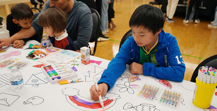 A boy draws a rainbow. At the table next to him, a man draws with two younger children leaning on him and watching him.