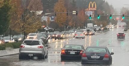 Vehicles traveling in high water on a flooded street. Additional vehicles and trees in the background.