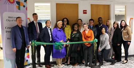 City and school district leaders stand in a room at the Welcome Center behind a green ribbon that they will cut.