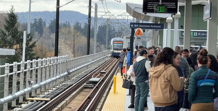 A crowd of people wait at a light rail station as a train arrives on the track.