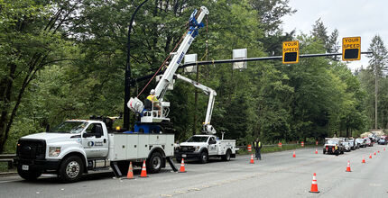 A boom truck and bucket truck with one worker each is erecting a large radar sign pole across a four-lane road. Traffic has been stopped by a uniformed police officer, and traffic cones are in place to divert traffic away from work crews.