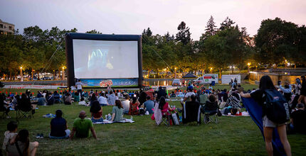 People sitting on the lawn at Downtown Park watch a movie on a massive, temporary screen.