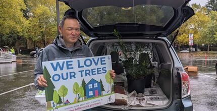A man holds a sign that says "We love our trees" while he stands in front of his car, which is loaded with free trees.