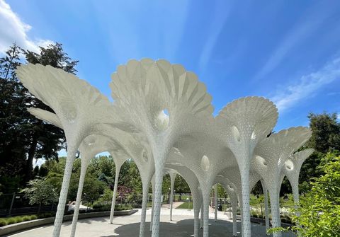 A photograph of a large sculpture. It is cluster of fluted columns rise up to form a canopy with perforated light coming through. In the background is Downtown Park.