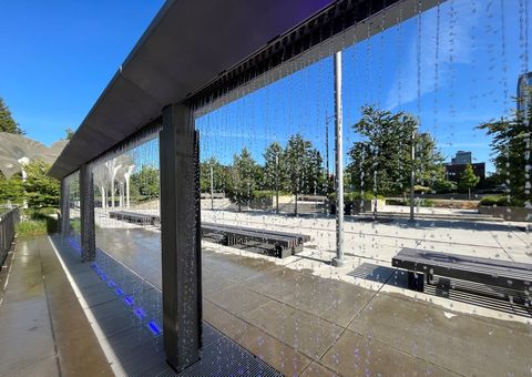In the foreground, water droplets fall in streams from a water feature. Behind this, several benches are seen in a concrete plaza. To the left, in the distance, you can see the white columns and canopy of the sculpture, Piloti.