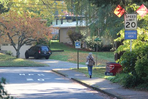 A photo of a residential neighborhood. In the foreground is a road leading to a T intersection. Just before the intersection, there are large white letters on the road that say "20 MPH". On the side of the road is a sidewalk. A person is walking on the sidewalk away from the camera. To the right of the sidewalk, there is a white "SPEED LIMIT 20" sign, with a blue "RESIDENTIAL AREA" sign below it. Both signs are mounted on a pole. At the top of the signs are two orange flags.