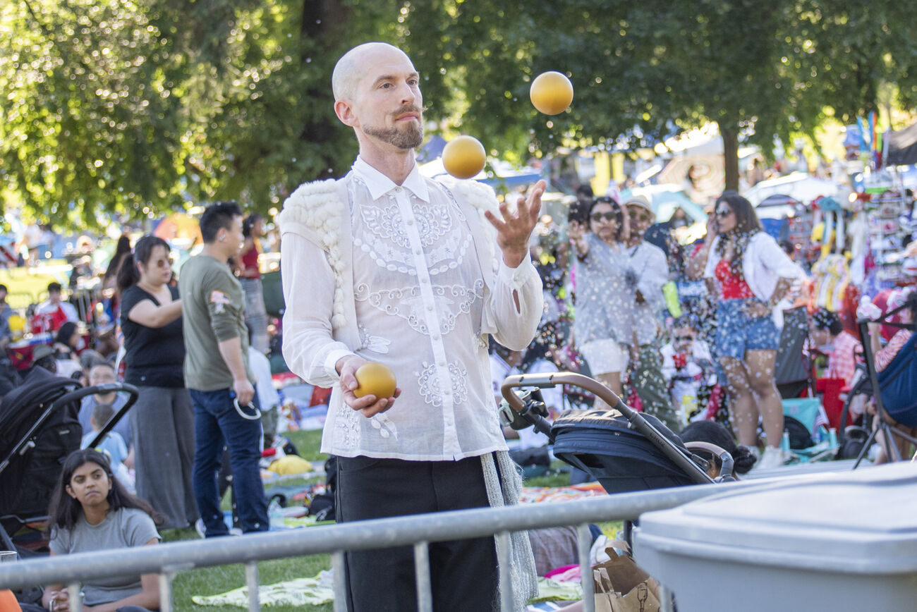 A man juggles three balls at Downtown Park while many people enjoy themselves behind him.