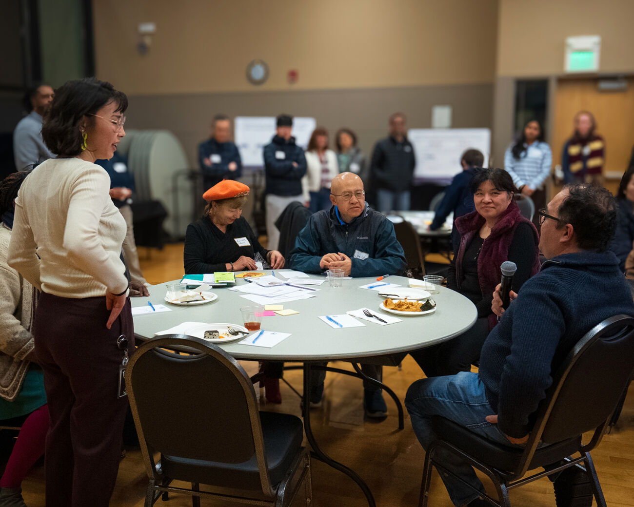 A man speaks into a microphone at a table with several people, in a large room with even more people.