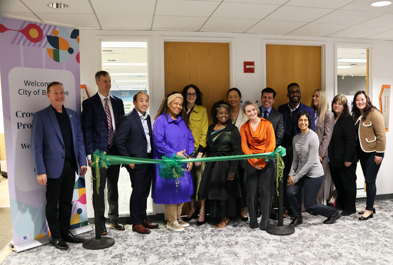 City and school district leaders stand in a room at the Welcome Center behind a green ribbon that they will cut.