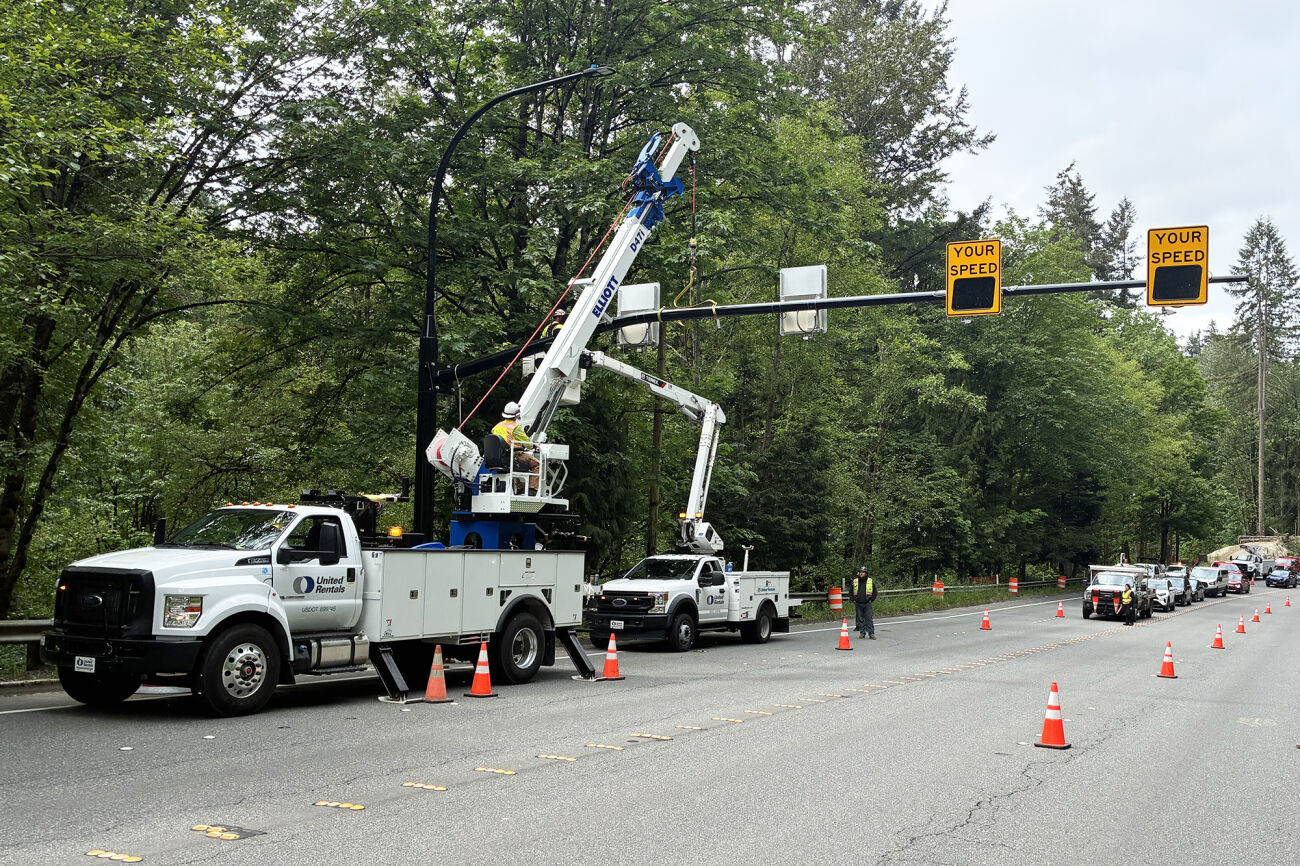 A boom truck and bucket truck with one worker each is erecting a large radar sign pole across a four-lane road. Traffic has been stopped by a uniformed police officer, and traffic cones are in place to divert traffic away from work crews.