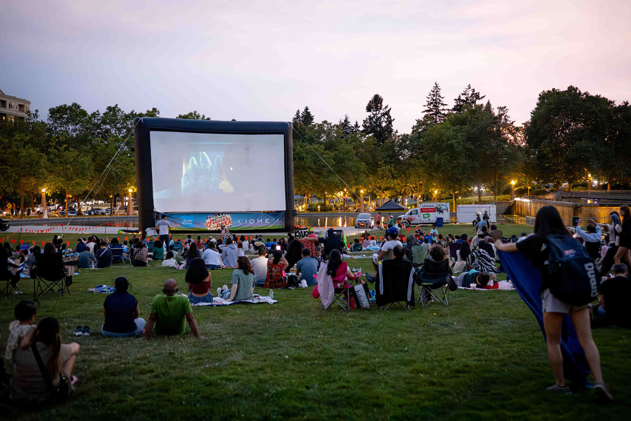 People sitting on the lawn at Downtown Park watch a movie on a massive, temporary screen.