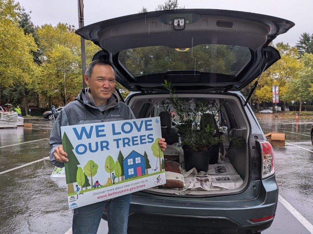 A man holds a sign that says "We love our trees" while he stands in front of his car, which is loaded with free trees.