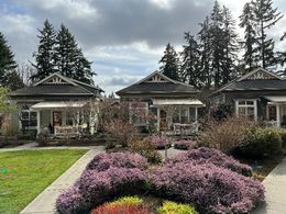 A picture containing grass, trees, outdoor sky and cottage style houses. There are three cottages with one story, front porches and walkways that lead to the entry. Shrubs with purple flowers line the paths as well as other plants. 