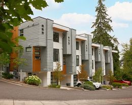 The photo shows townhomes attached next to each other with driveways in front of garage doors. The townhomes are three stories with flat roofs and there are 6 side by side. They feature green landscaping with flowers and trees. 