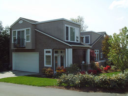 Image shows an example of a detached accessory dwelling unit over a garage. The image shows a two story detached accessory dwelling unit with grey shingles and white doors and trim. There is a driveway that leads to a garage door. The image also shows green grass with landscaping of shrubs and flowering plants and trees behind the home. 