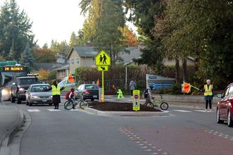 Two crossing guards stop traffic at a crosswalk. Two children walking bikes are using the crosswalk to cross the road.
