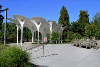 In the foreground, there is a plaza with greenery and benches. In the background, the fluted columns of the sculpture Piloti rise up to form an undulating canopy.
