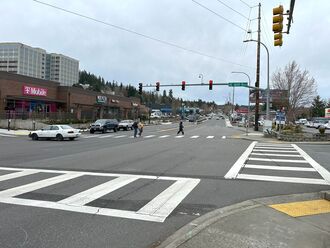 An intersection with shops on both sides of the street and offices in the background. People are crossing the street in a crosswalk and there are cars driving through the intersection parallel to the people.
