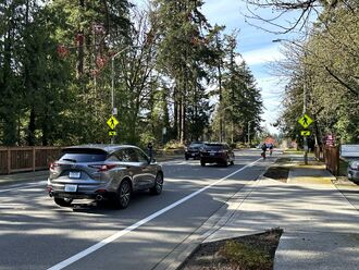 A two lane road with cars driving on both sides. There is a striped bike lane on each side, and people are cycling away from the viewer. There is also a flashing pedestrian beacon.