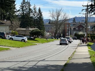 A neighborhood street with cars driving away from the viewer. There is a group of people walking on the sidewalk next to the cars; the other side of the street has no sidewalk.