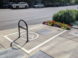 White box painting on the sidewalk with a bike rack in the middle and a flower planter on the right side. 