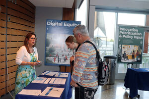 Three people converse at an information table, with the "Digital Equity" sign in the background.