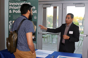 Two people converse at an information table, with the "Policy and Prioritization" sign in the background.