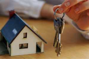 Hand holds set of keys next to a miniature house