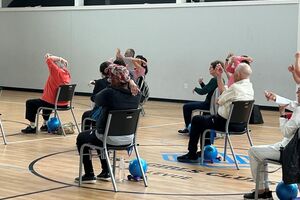 A group of people in a 55+ fitness class, sitting in chairs in various stretching poses